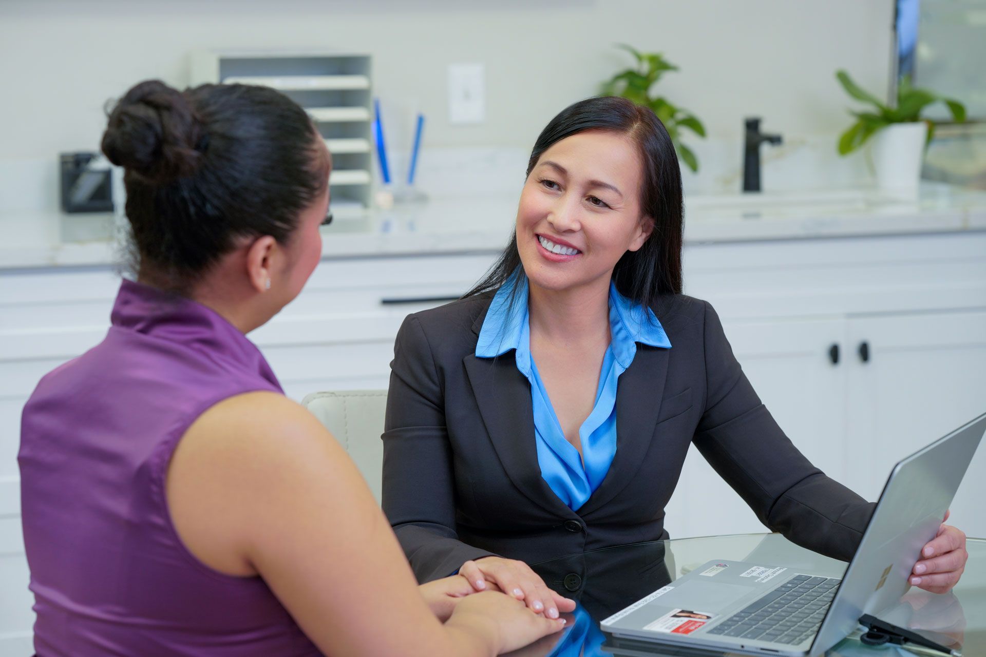 Woman in blazer smiles at a client sitting across a table, both facing a laptop in a brightly lit office.