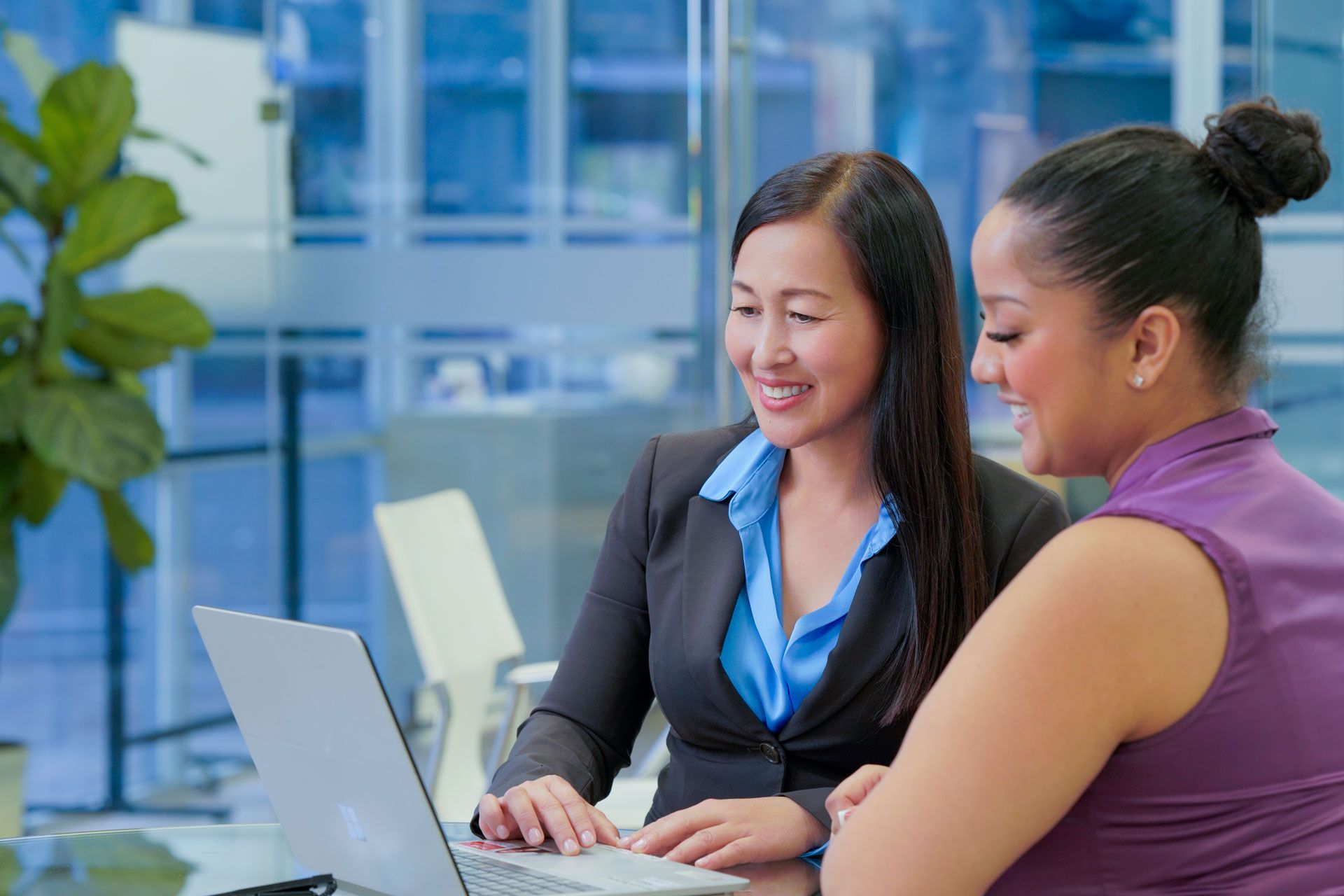 Two women smiling at a laptop in a modern office, one typing.