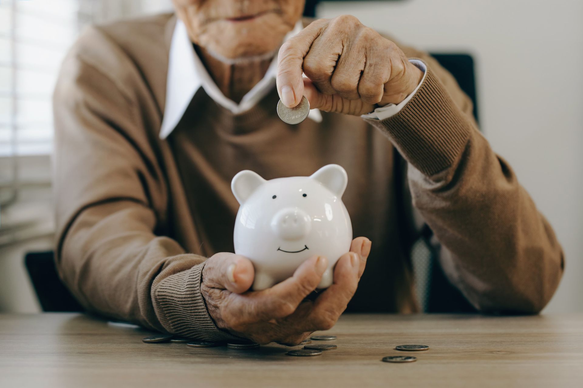 An elderly person putting a coin into a piggy bank, sitting at a table.