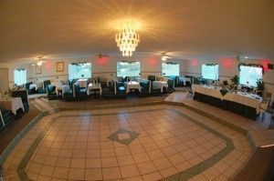 Wide banquet hall with tiled floor, chandeliers, and rows of tables by windows and columns.