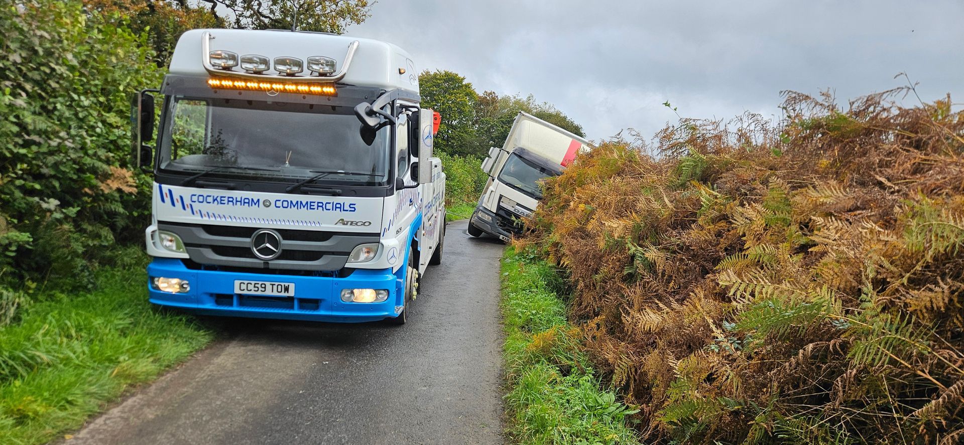 Blue coach bus tilted on a narrow rural road, with another vehicle behind it and overgrown hedges beside the lane