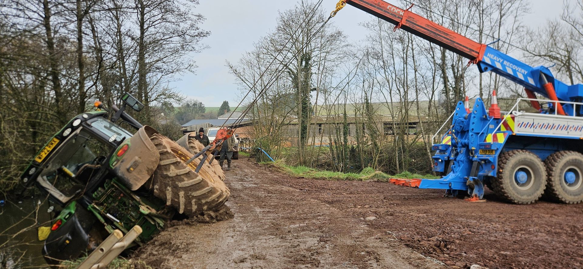 Overturned tractor beside muddy road as a crane lifts it upright near a wooded field
