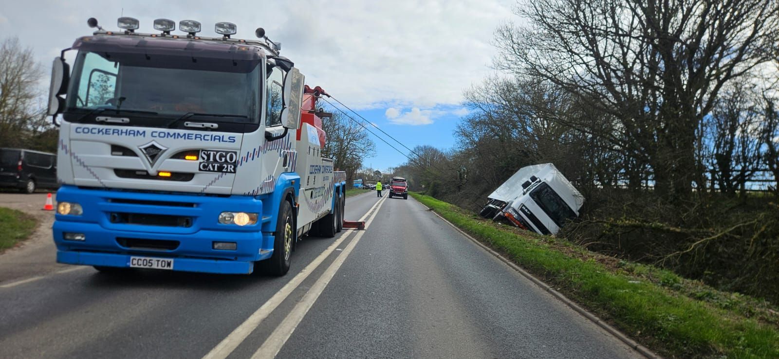Blue tow truck on a rural road beside a crashed white van in a ditch