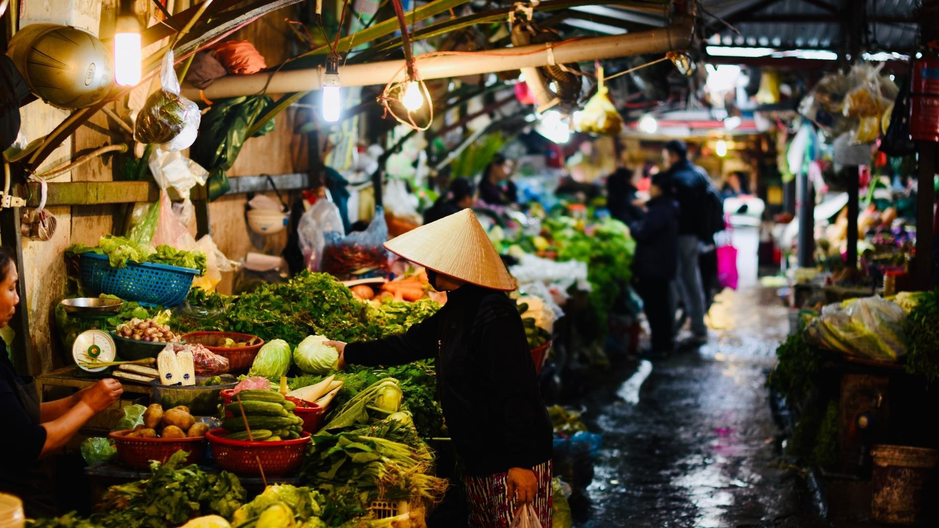 A woman in a conical hat is selling vegetables at a market.