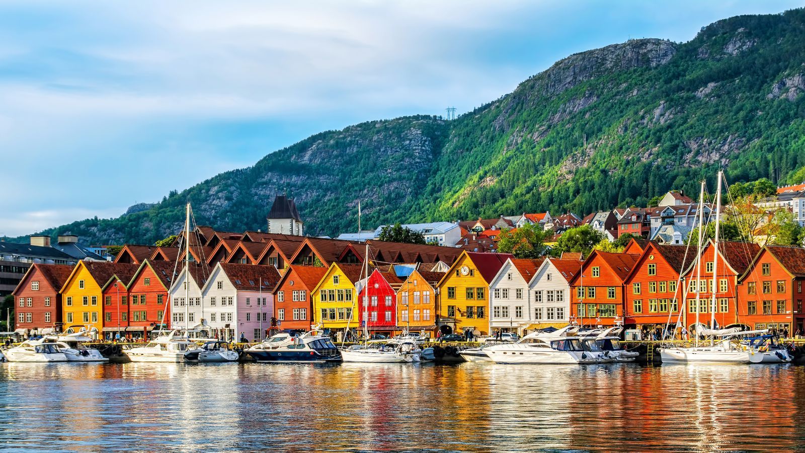 Colorful wooden buildings line a harbor in Bergen, Norway, with boats and a green mountain backdrop.