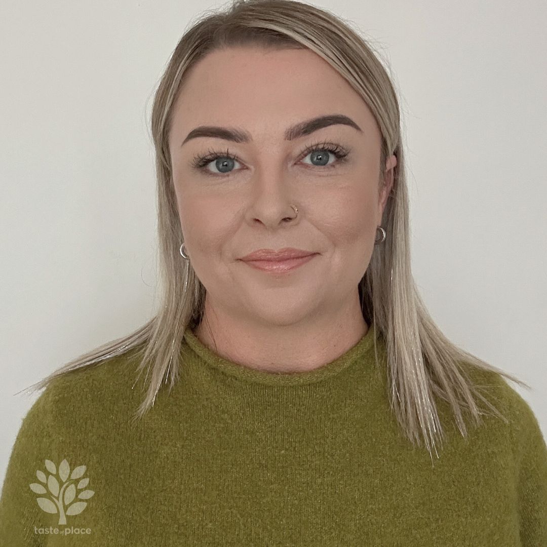 Woman with blonde hair wearing a green sweater smiles at the camera against a white wall.