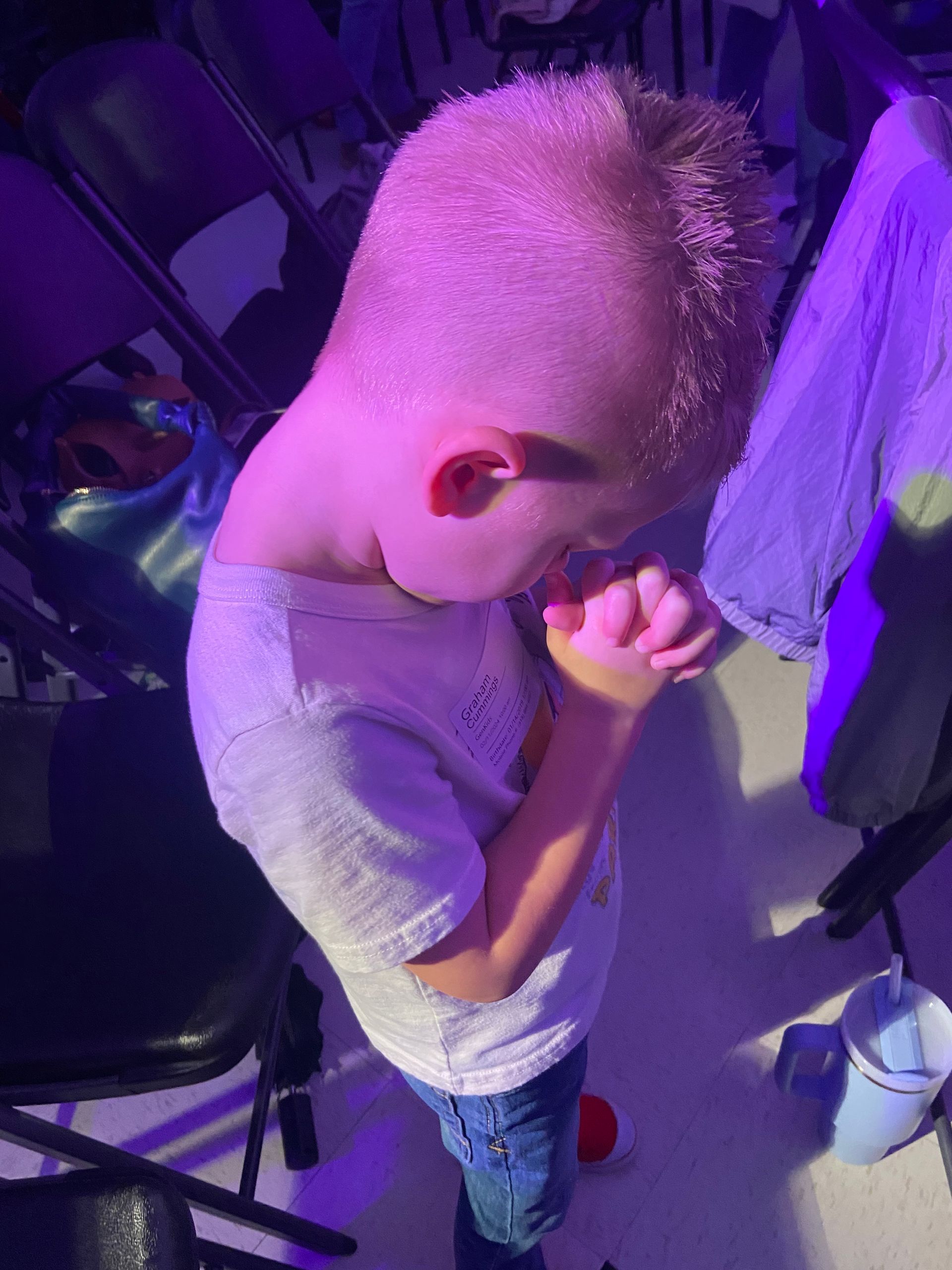 A young boy is praying with his hands folded in front of a purple light.