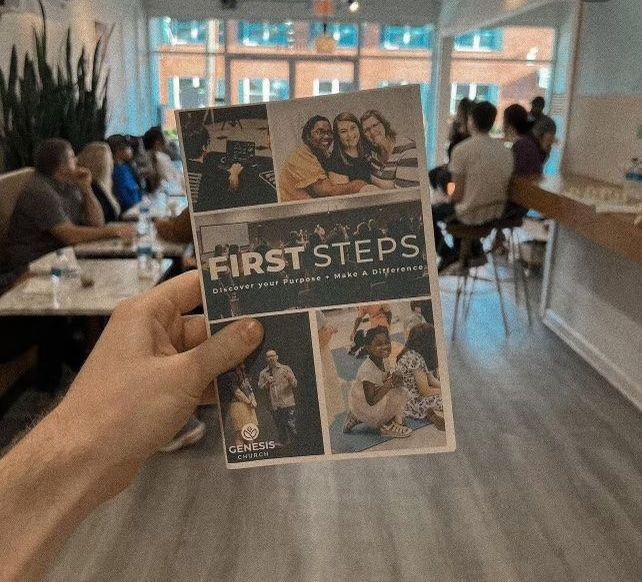 Hand holding a “First Steps” brochure in a bright waiting room with seated people and plants.