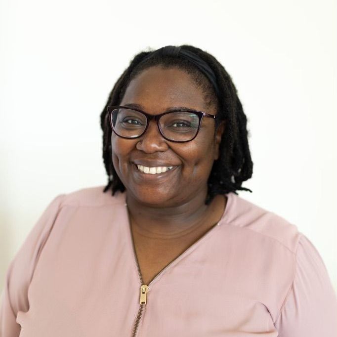 Woman with glasses smiles; wearing a pink shirt, in front of a white background.