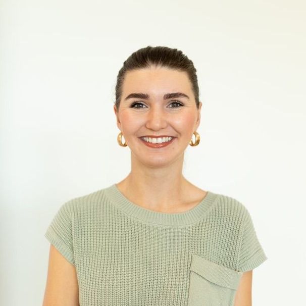 Woman with brown hair in a sage green sweater smiles, wearing gold hoop earrings, in front of a white wall.