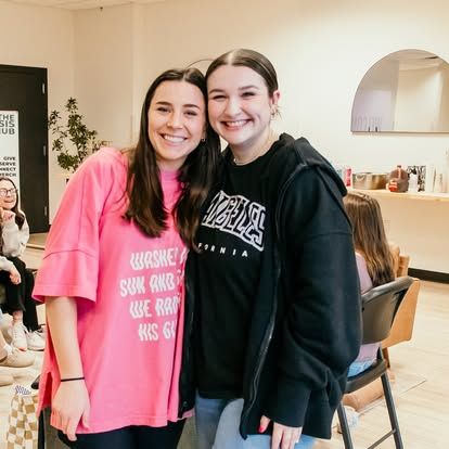 Two smiling people posing indoors, one in a pink shirt and one in a black hoodie.