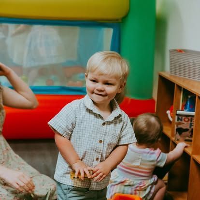 Smiling toddler in a plaid shirt standing in a colorful play area with another child nearby