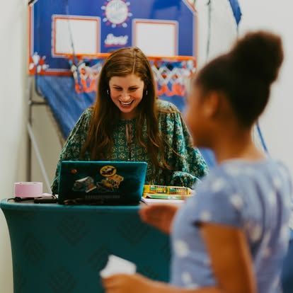 Woman smiling behind laptop at table during a game with a child in front of a basketball hoop backdrop