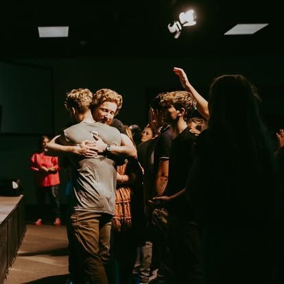 People hugging in a dimly lit room during a small gathering, with others standing nearby.