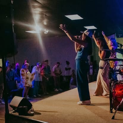 People worshipping on a stage in a dim church hall, with raised hands and bright ceiling lights