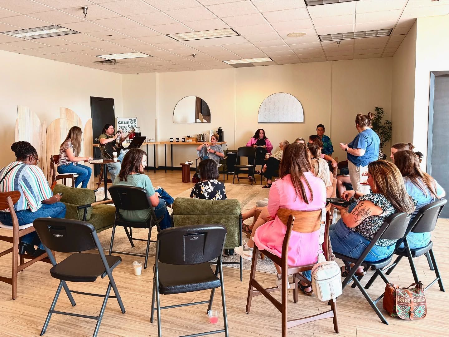 People seated in a circle in a bright room, listening to a speaker at the front.