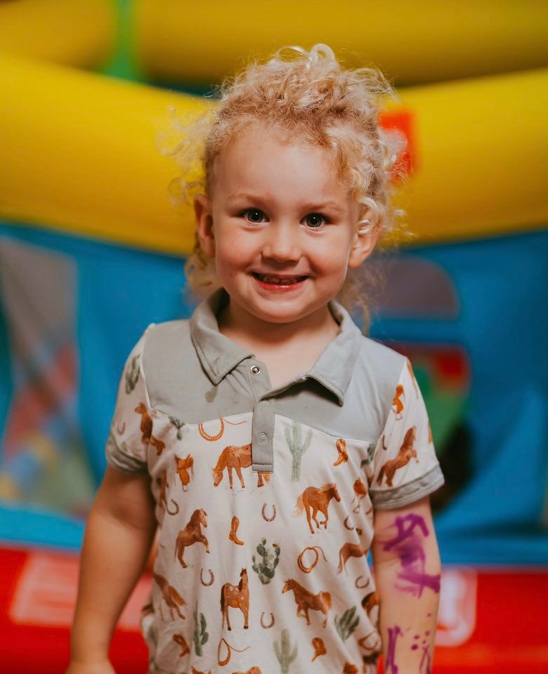 Smiling child standing indoors in front of colorful inflatable play equipment, wearing a patterned shirt
