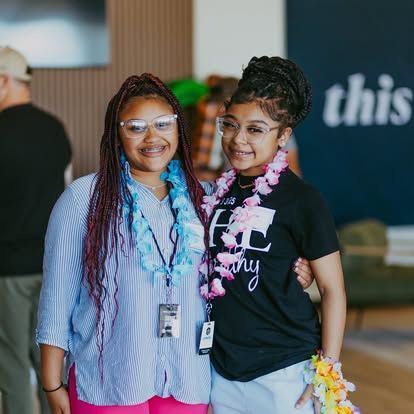 Two smiling people stand together indoors, wearing glasses and lanyards, in front of a blurred backdrop.