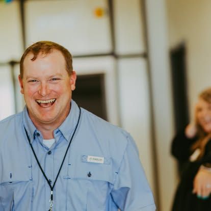 Smiling man in a light blue shirt at an indoor event, with a blurred woman in the background