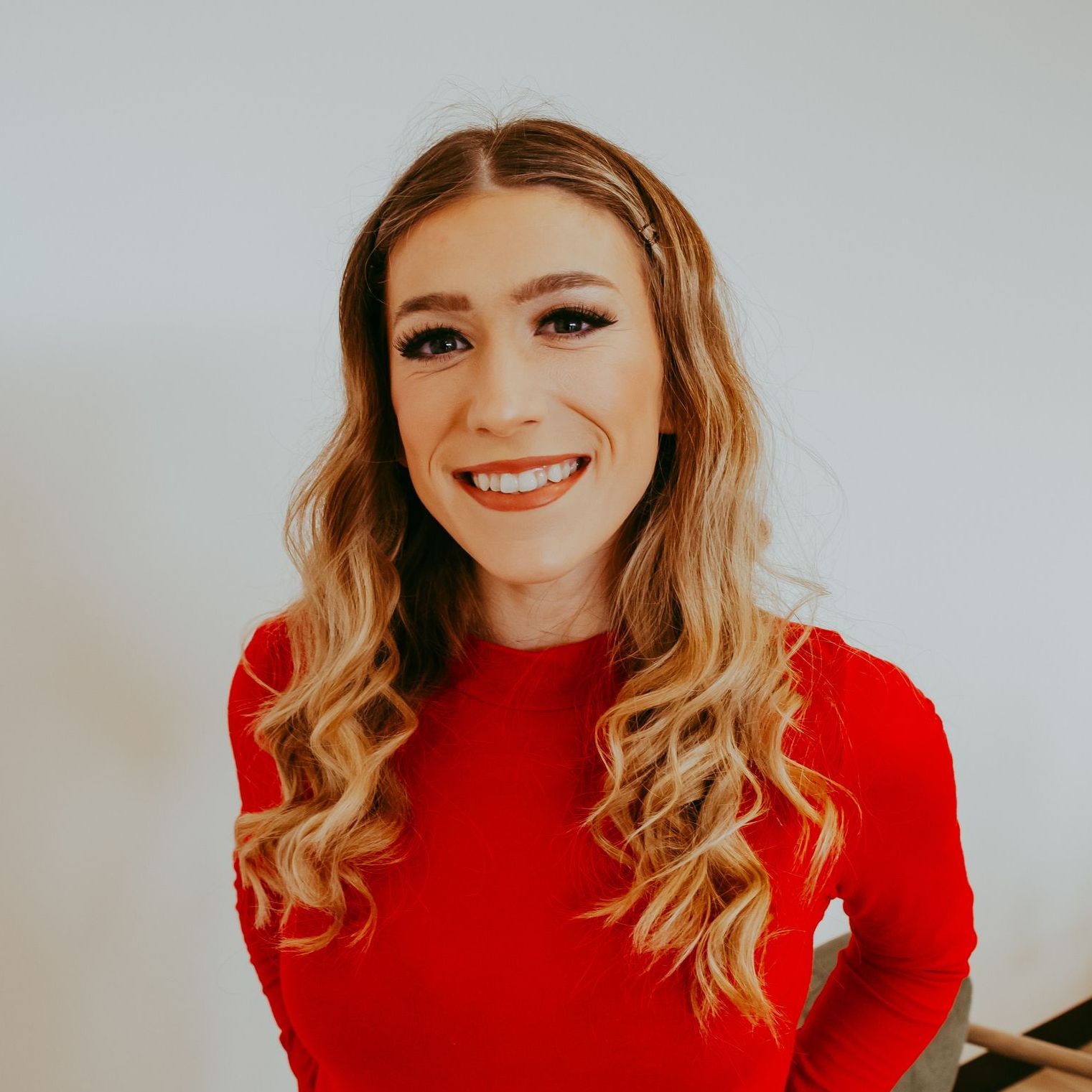 Woman with long, wavy blonde hair smiles, wearing a red long-sleeved shirt, facing the camera.