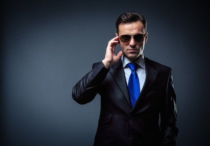Man in black suit and blue tie adjusting sunglasses against a dark studio background