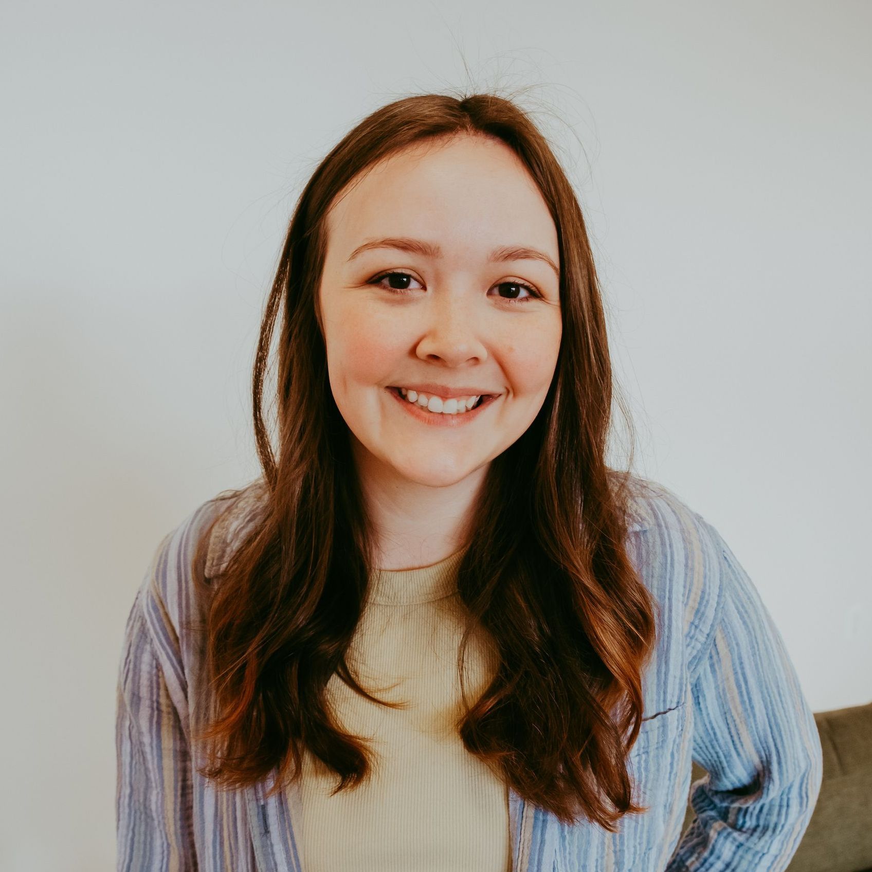 Woman smiling, brown hair, wearing a striped blue shirt over a yellow sweater, against a white background.