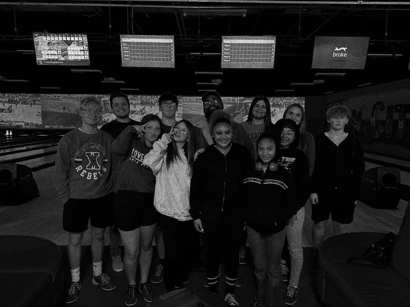 Group of people smiling at a bowling alley, scoreboard visible.