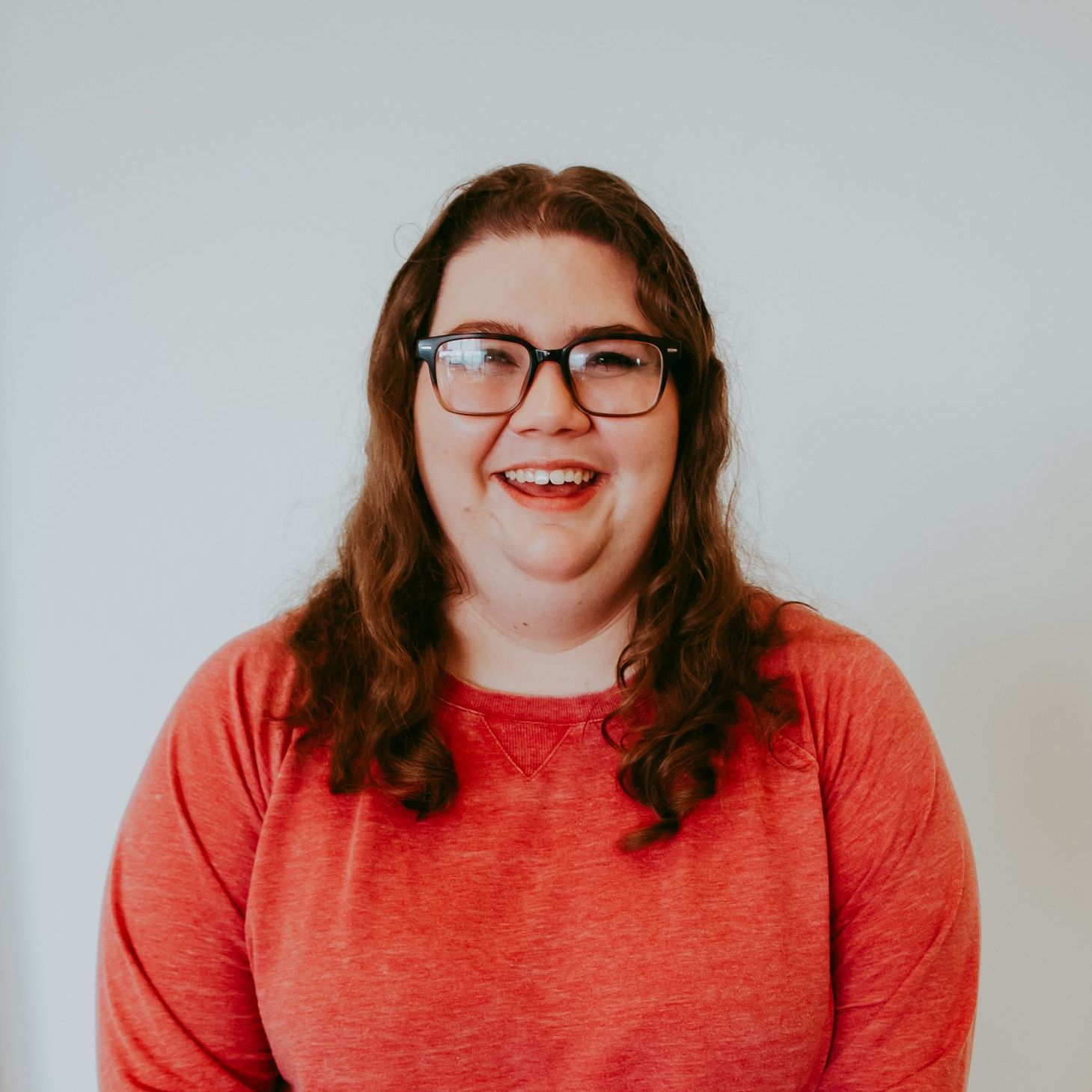 Woman with glasses and red shirt smiling in front of a white wall.