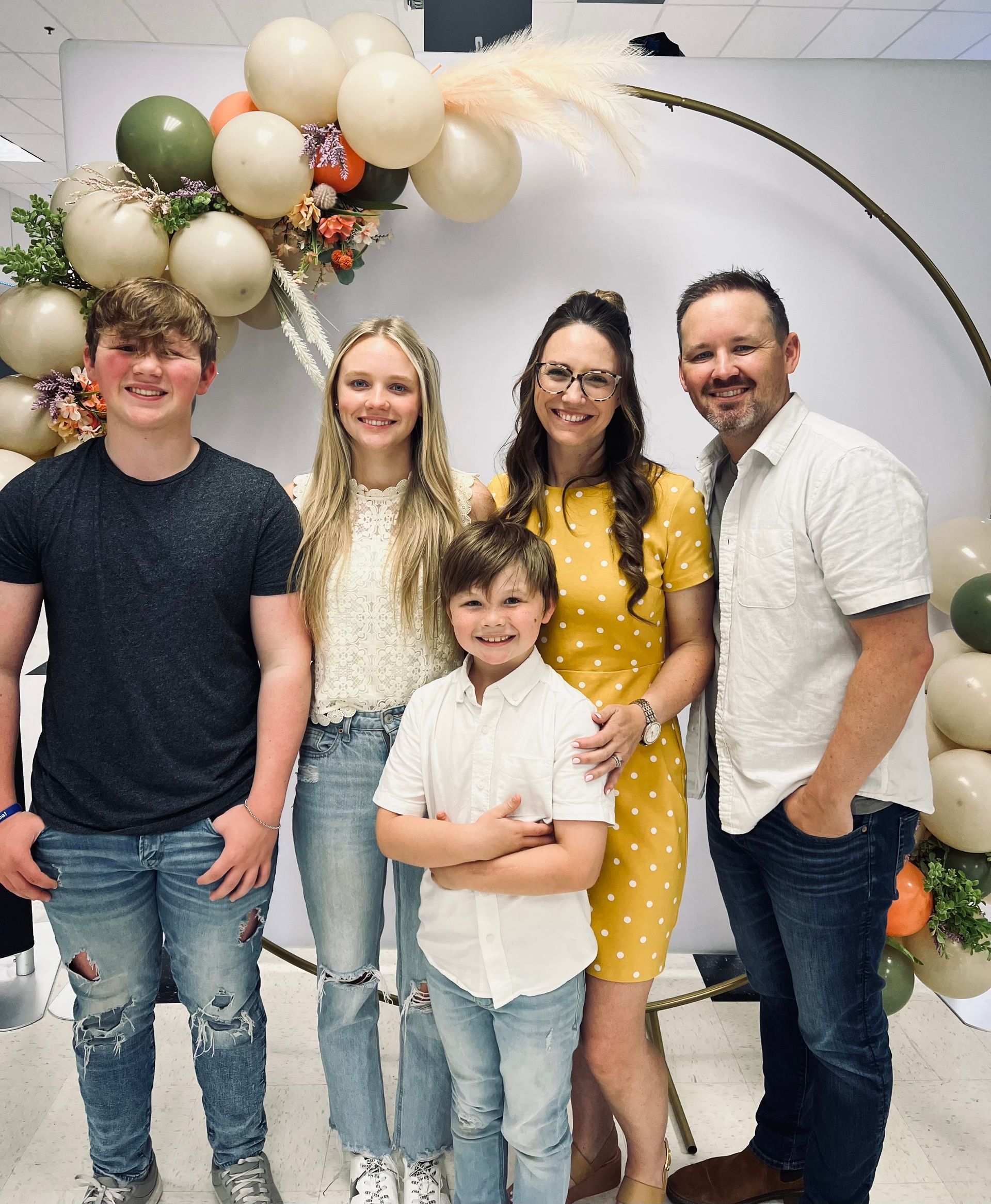 Family of five posing in front of a balloon arch; adults smiling, children with various expressions, neutral background.