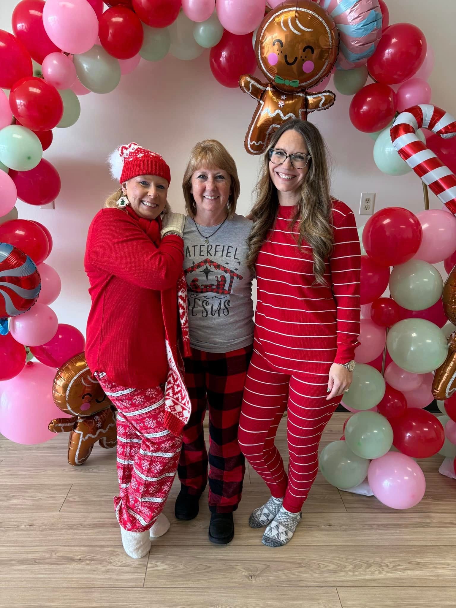Three people posing in festive red pajamas before a pink and red balloon arch, smiling at the camera.