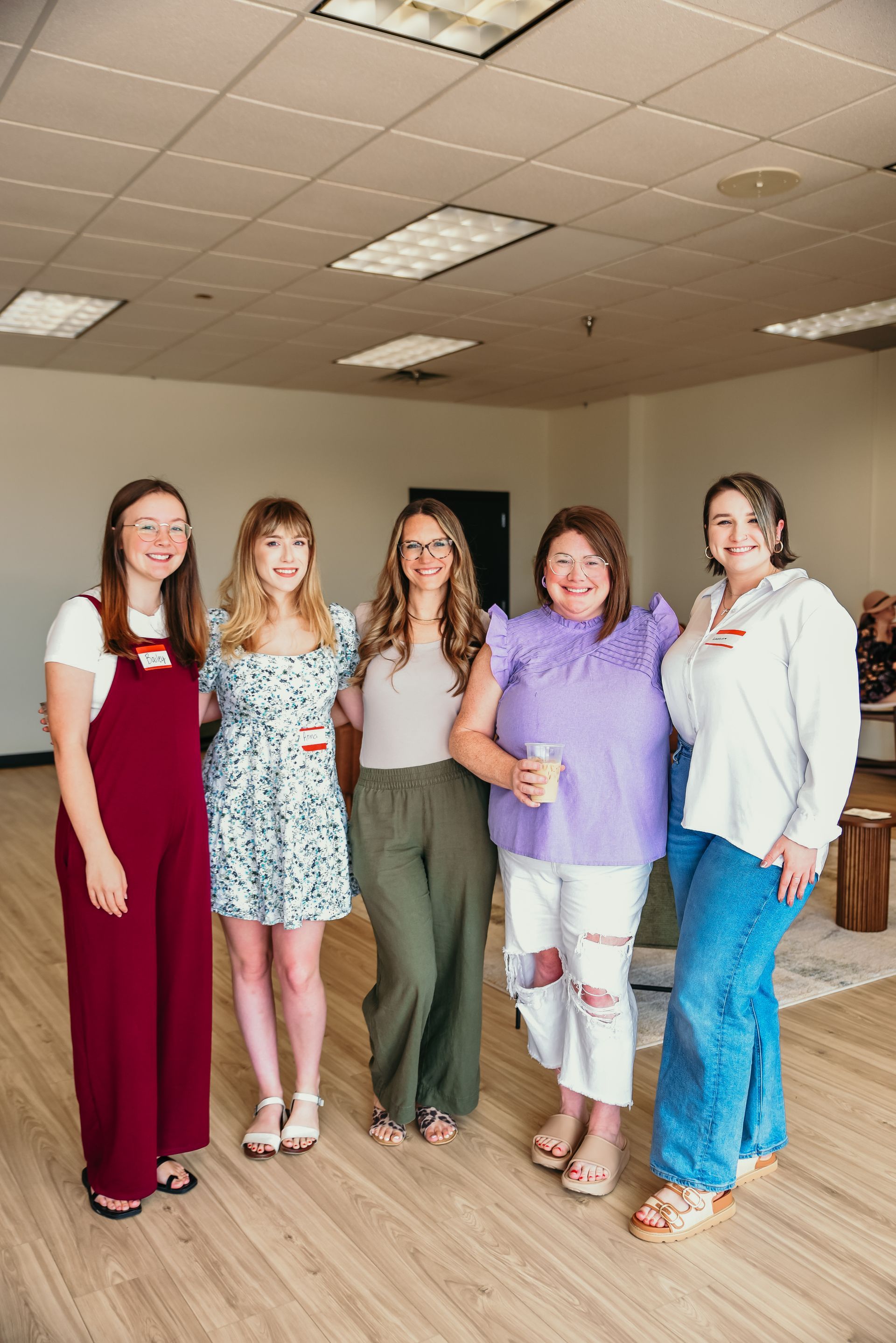Five people smiling, standing indoors. One in maroon overalls, one in a floral dress, another in green pants, a fourth in a purple shirt, and a final person in a white top and blue jeans.