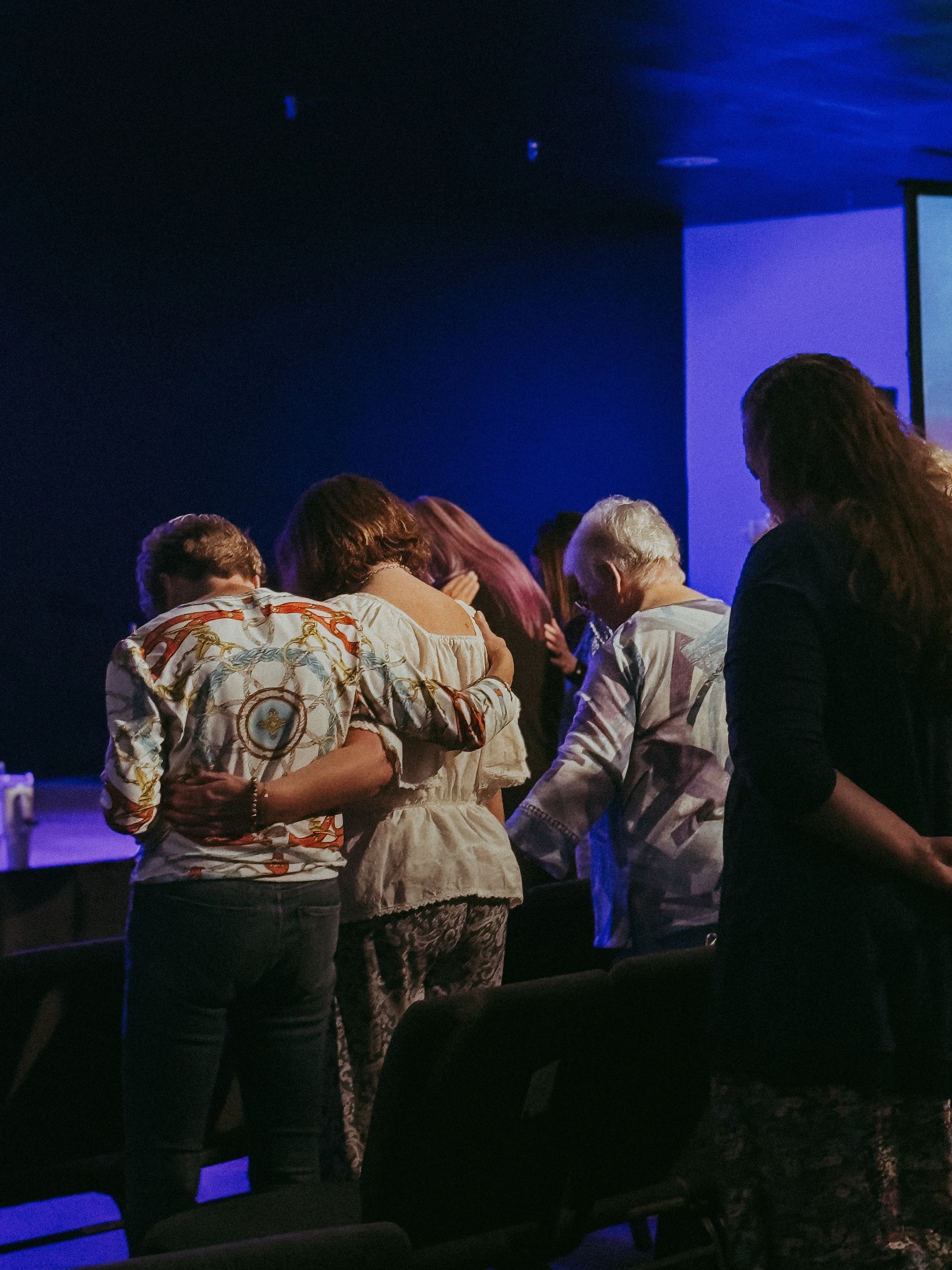 People praying together, some with arms around each other, in a dimly lit room, presumably a church.