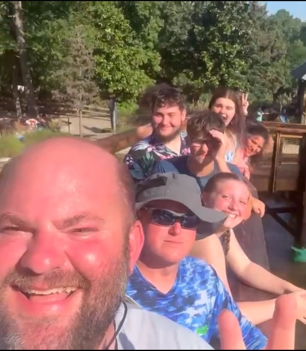 A group of people smiling for a selfie near a body of water and trees.