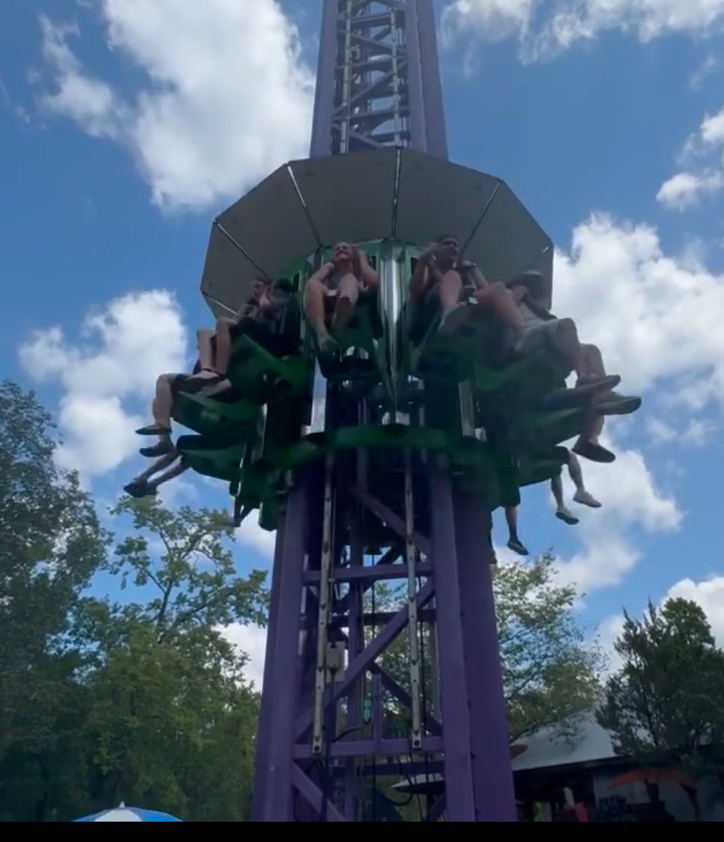 Tower ride with riders in green seats, ascending purple tower against a cloudy sky.