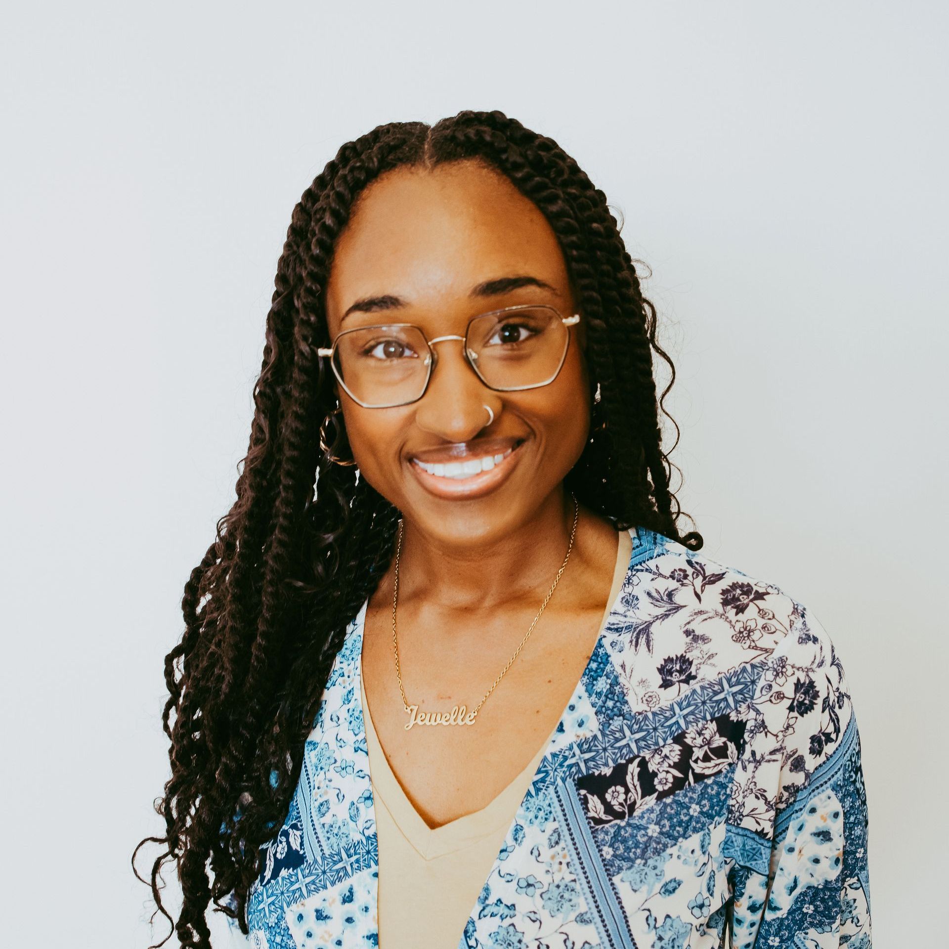 Woman with glasses smiles; wearing patterned blue top, gold necklace.