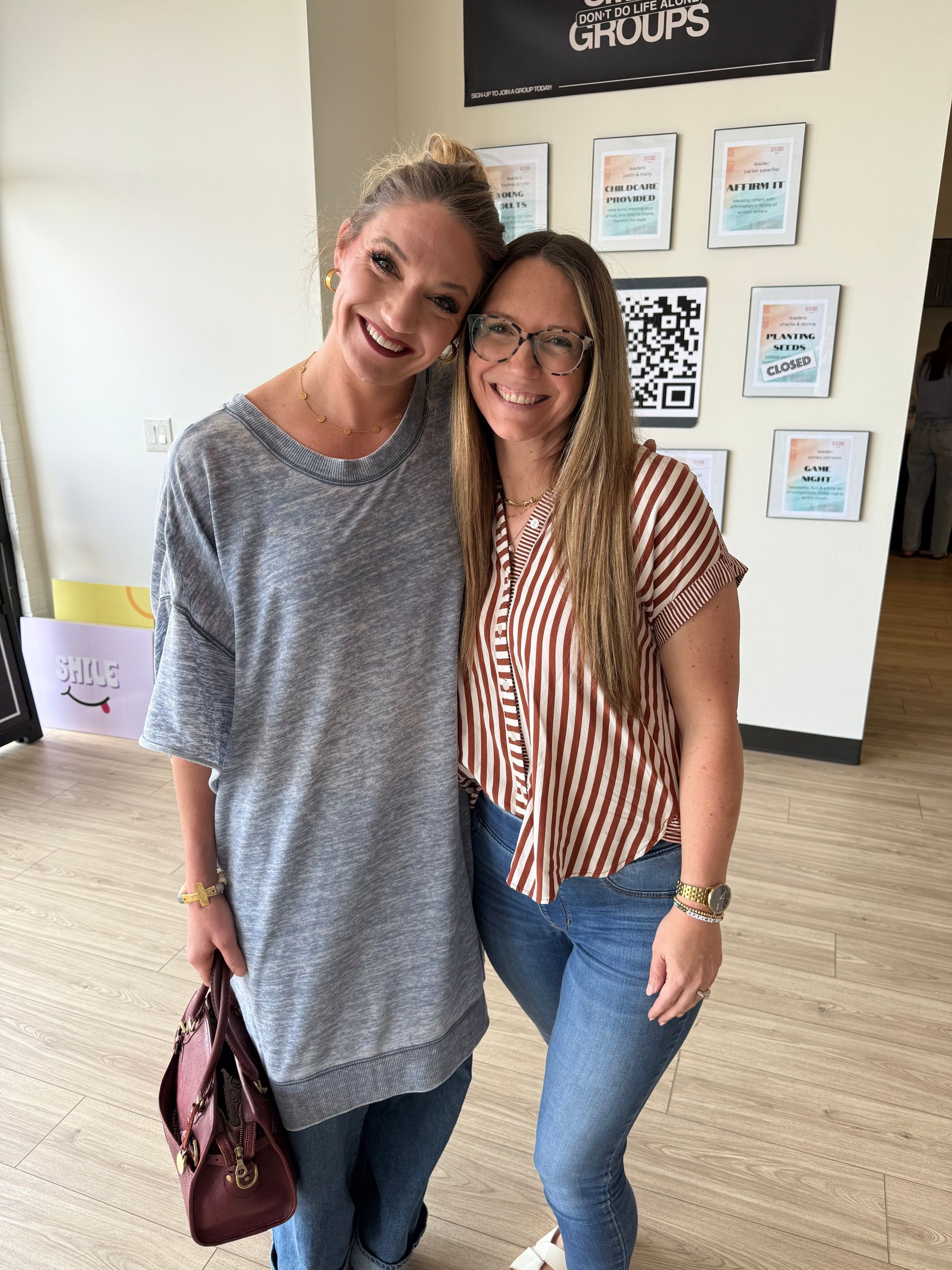 Two women smiling, arm in arm. One in a gray tunic, the other in a striped shirt, both wearing jeans. Indoors.
