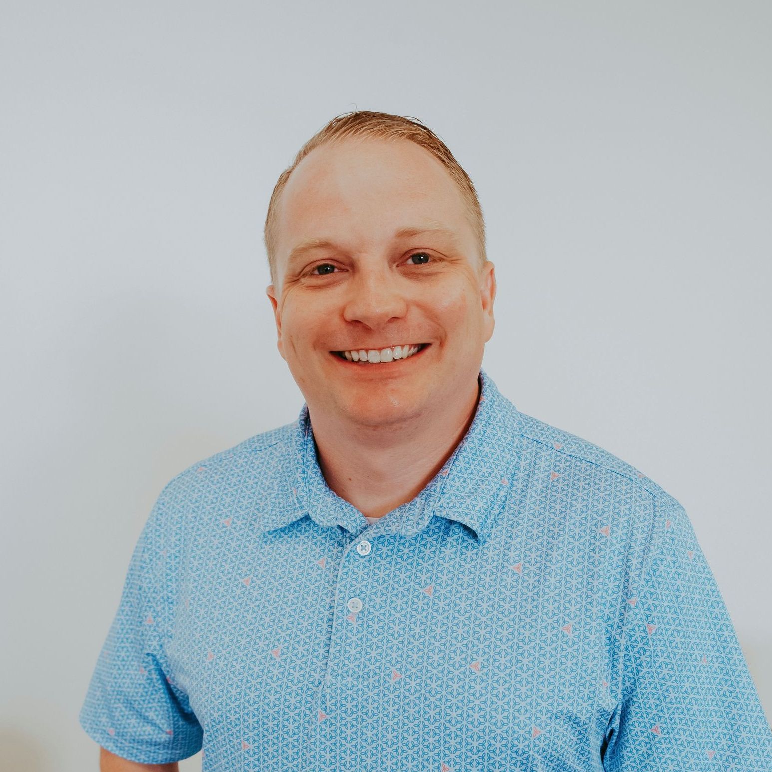 Man with fair skin wearing a blue patterned polo shirt smiles against a white background.