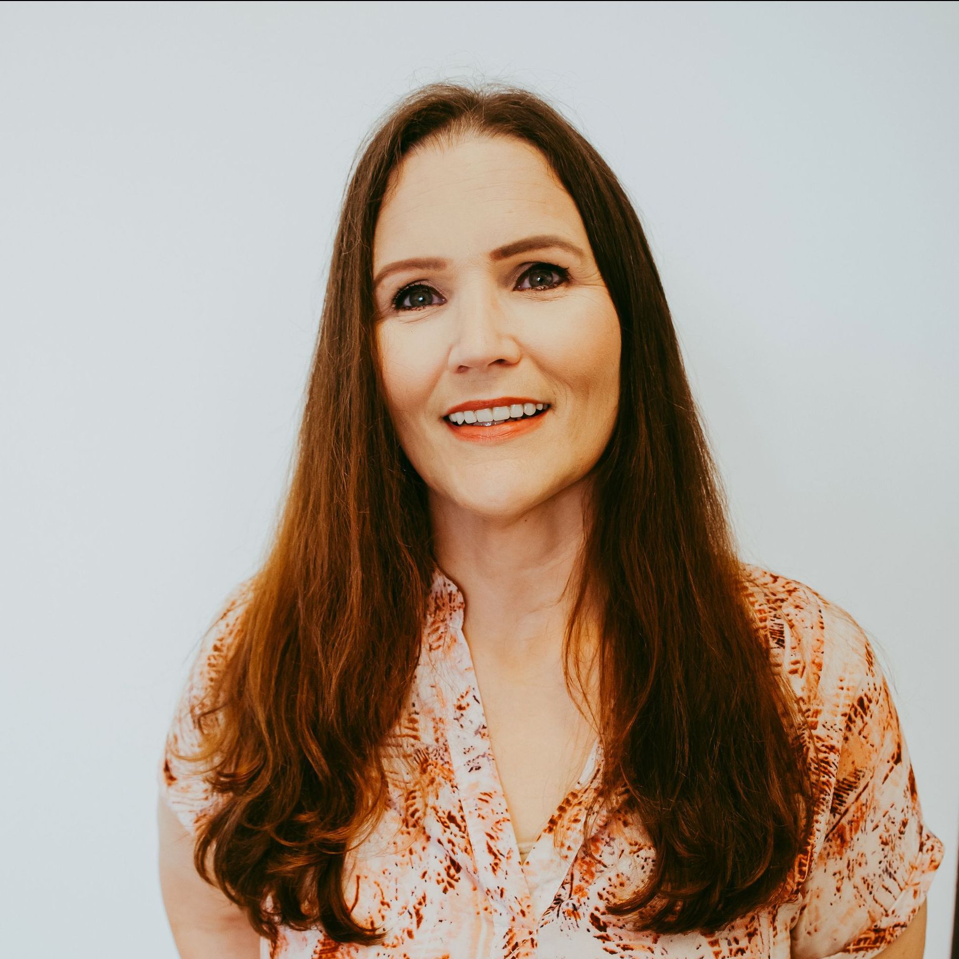 Woman with long brown hair smiles, wearing a pink patterned shirt, against a white backdrop.