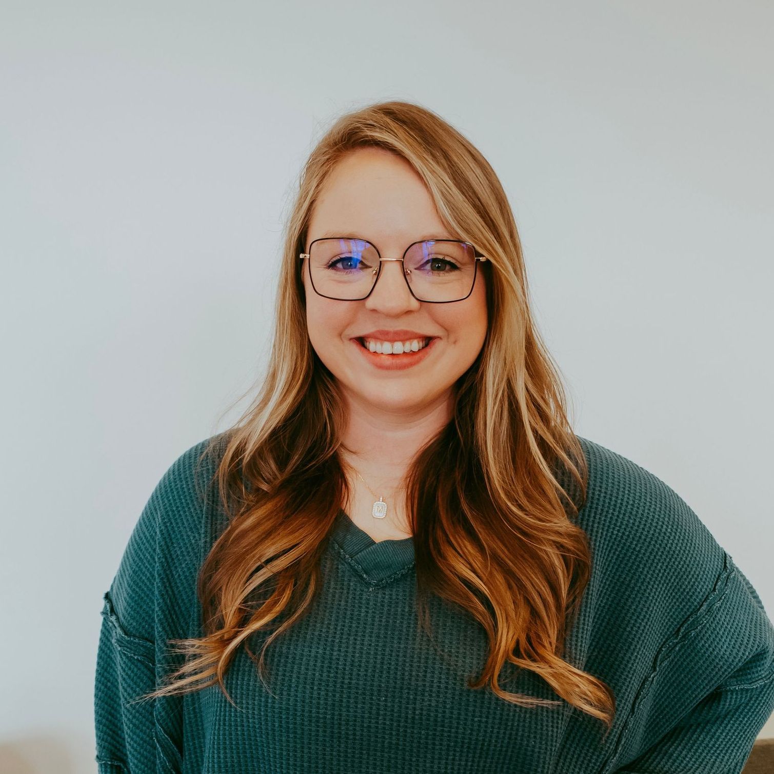 Woman with long, wavy blonde hair, wearing glasses and a teal sweater, smiling in front of a white wall.