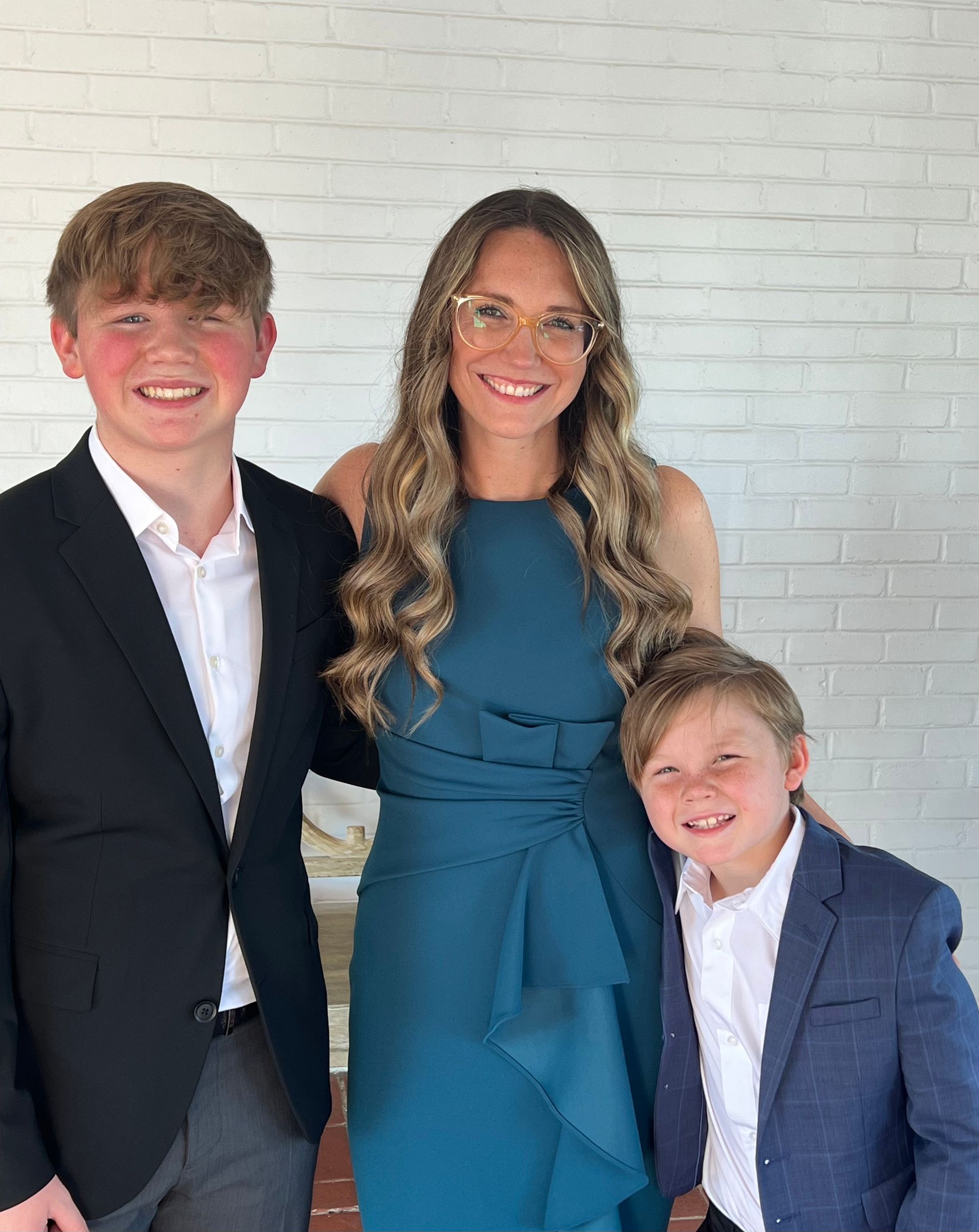 Woman in teal dress smiles with two boys, one in a suit. White brick background.