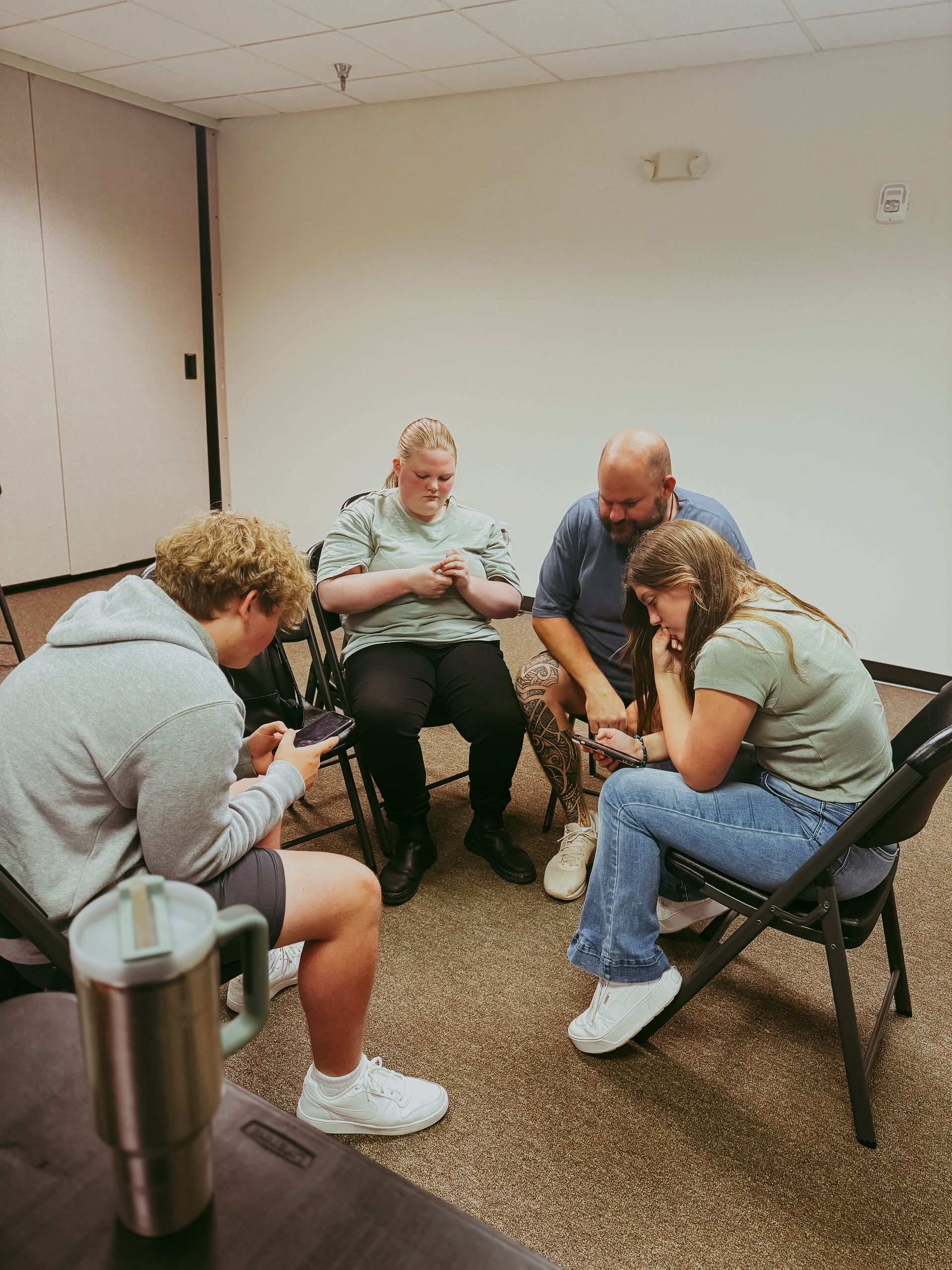Four people in a circle, hands clasped. One uses a phone. Neutral setting, carpet. Expressions of concern.