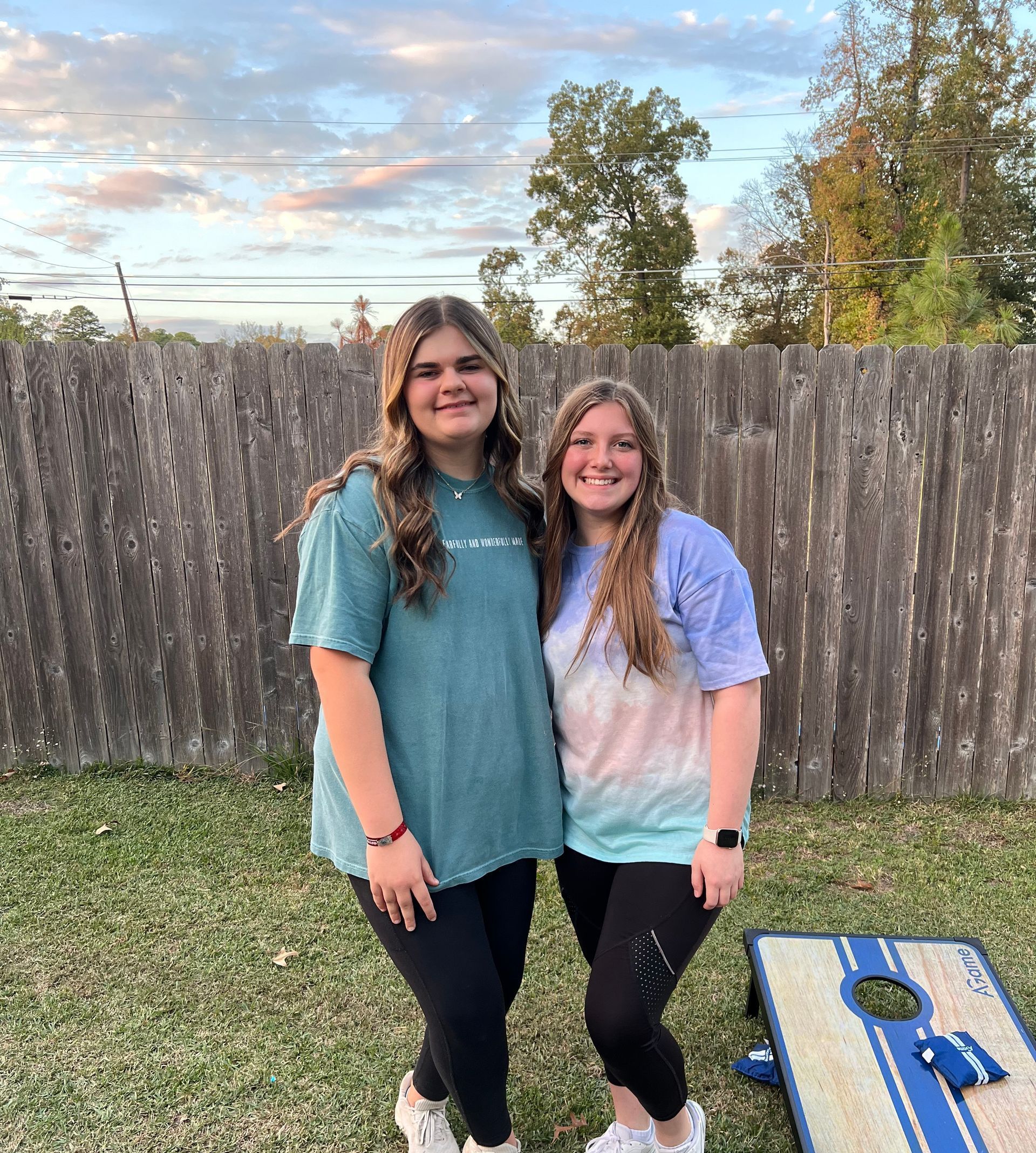 Two women standing next to each other on grass, near a fence, with a cornhole board in the foreground.
