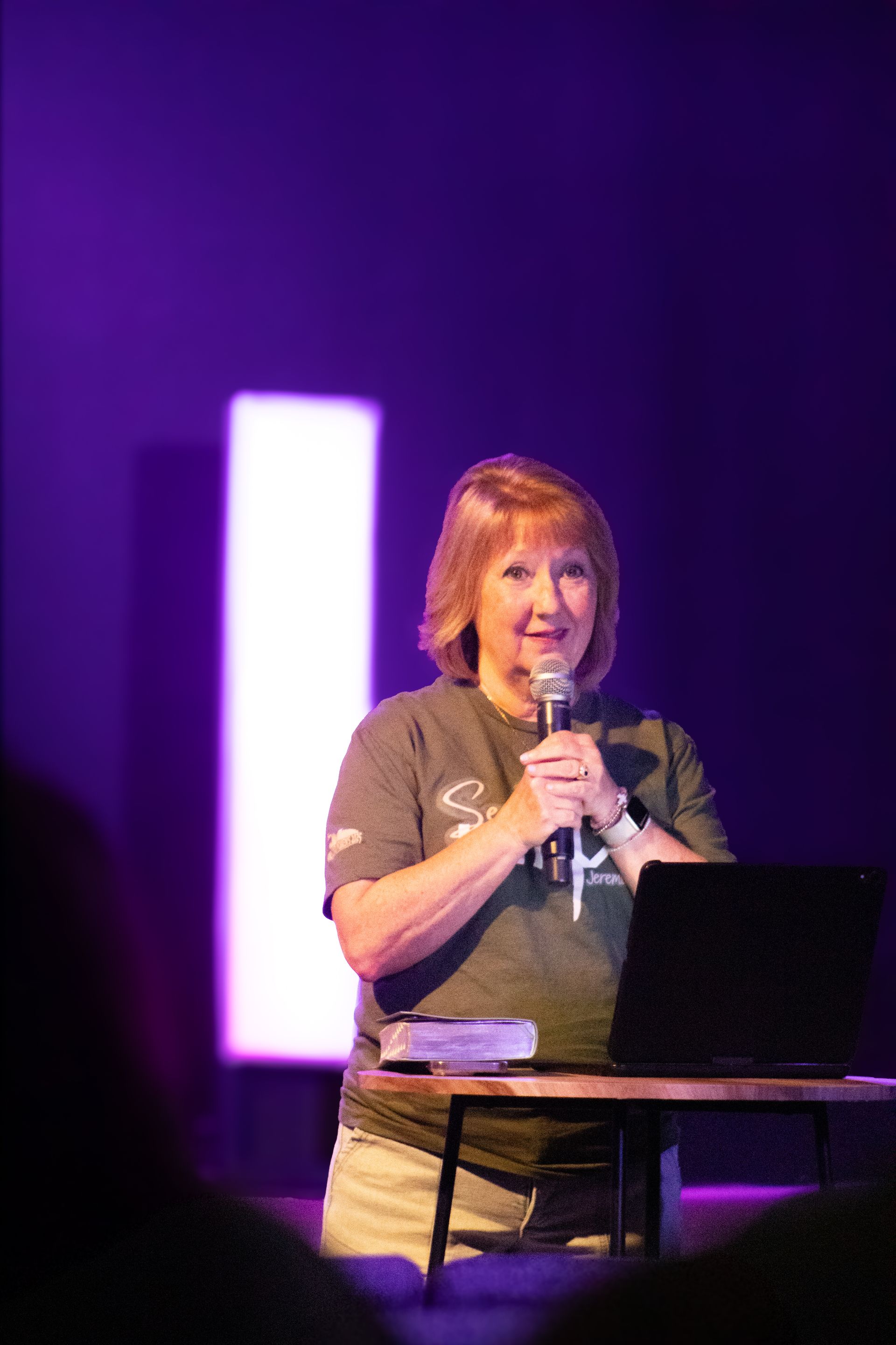 Woman speaking into a microphone on a stage with purple lighting and a laptop on a table.