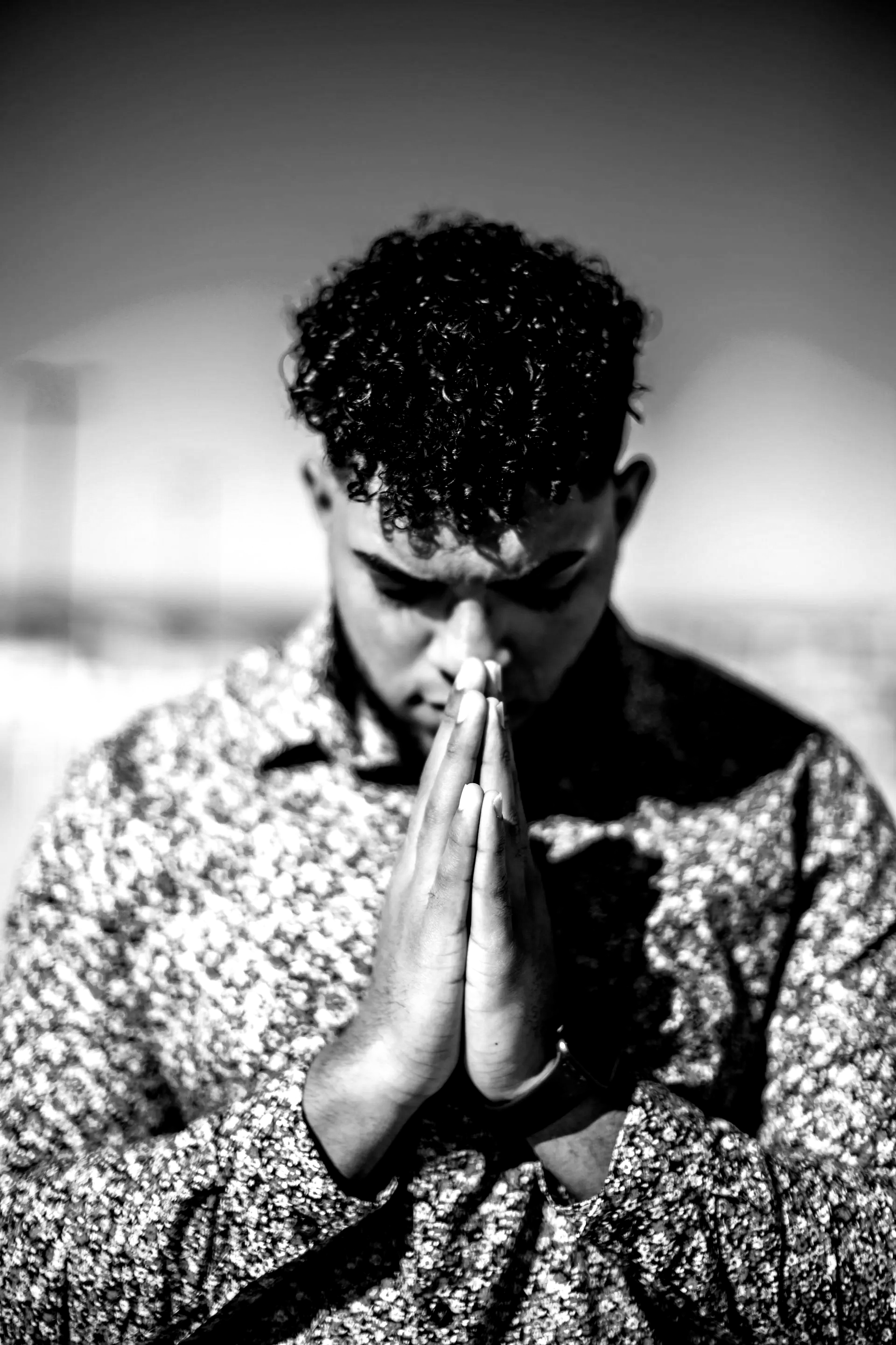 Man with curly hair, hands clasped in prayer, eyes closed, wearing floral shirt.