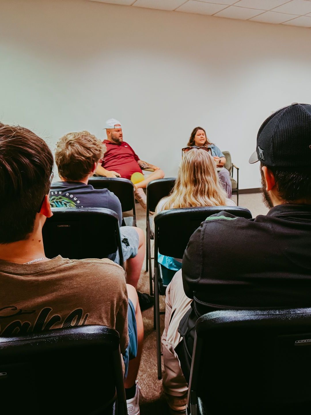 People seated, facing a panel of two, in a room with a white wall.