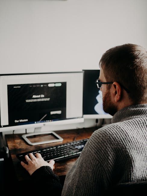 Une personne tapant au clavier à un bureau équipé de deux écrans dans un espace de travail faiblement éclairé