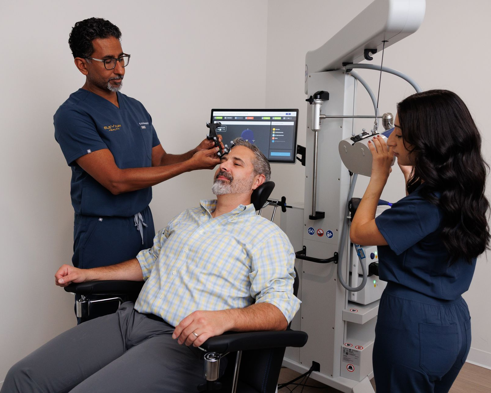 Patient undergoing eye exam with clinician using equipment and another assistant beside a monitor