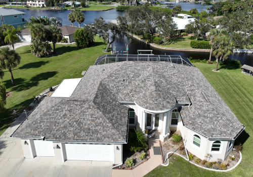 Aerial view of a gray-roofed house with a waterfront view, surrounded by green grass and trees.