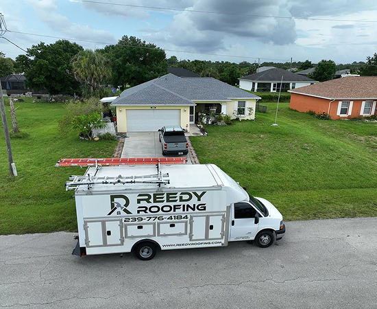 White Reedy Roofing truck parked in front of a yellow house with gray roof; another house in the background.