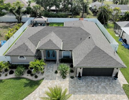 Aerial view of a gray-roofed house with landscaping and a driveway; white fence surrounds the property.